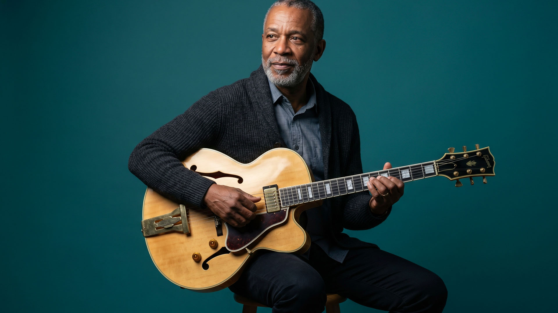 Jazz guitarist holding an archtop guitar against a deep teal studio backdrop